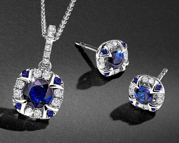 A necklace and earrings of sapphire set in a double diamond halo in white gold on a black textured background.