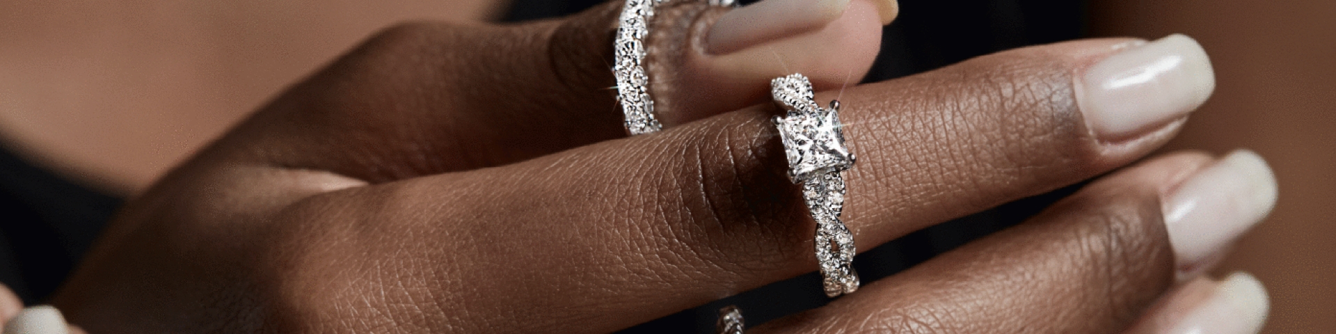 Woman's hand holding a white gold diamond engagement ring with a princess cut center stone on a black background.